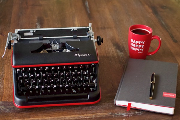 A typewriter, notebook, pen, and coffee cup sitting on a wooden table.